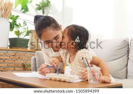 Asian cute daughter kissing her mother cheek while playing and painting ceramic models on the table using colorful watercolor in living room at home Happy family and mother and child lifestyle concept