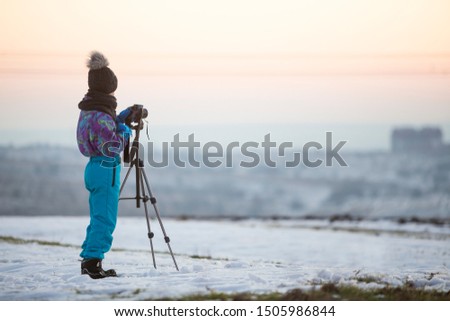 Child boy taking pictures outside in winter with photo camera on a tripod on snow covered field.