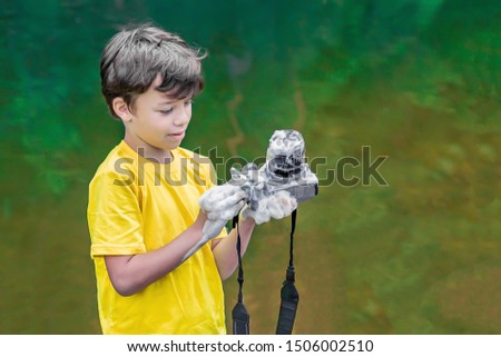 A child in a yellow t-shirt and shorts is standing near the water in nature and washing a camera with soap and foam. The concept photographers day.