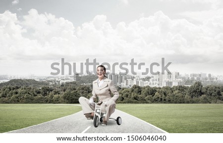 Beautiful happy woman riding children's bicycle on asphalt road. Young employee in white business suit cycling small bike outdoor. Nature landscape with green grass. Professional career start concept