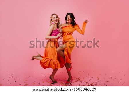 Two attractive girls dancing at party. Indoor full length photo of carefree female models posing on pink background.