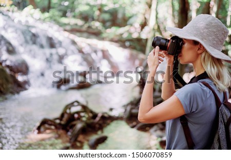 Travel and technology. Young woman in hat with rucksack taking photo of  waterfall in jungle.