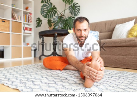 Young attractive nice man do yoga practice at home near grey sofa on carpet. Sitting relax meditative asana near couch. Basic yoga workout that helps to fit body and mind.  Male  white t-shirt 