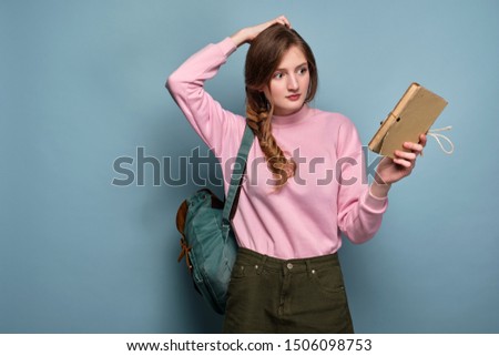 A girl in a pink sweater with a backpack stands on a blue background and holds a book, looking puzzled at her, scratching her head