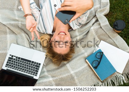 Top view of a beautiful young blonde girl relaxing on a lawn at the park, taking a selfie while laying on a blanket with laptop computer