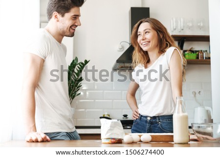 Image of happy young couple rejoicing while cooking breakfast together with milk and eggs in apartment
