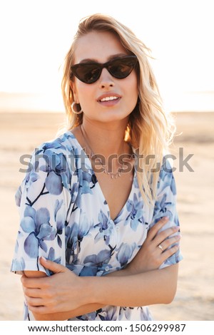Photo of seductive young woman wearing sunglasses looking at camera with arms crossed while walking on sunny beach