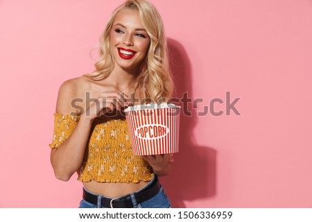 Image of adorable woman with red lipstick smiling and holding popcorn bucket isolated over pink background