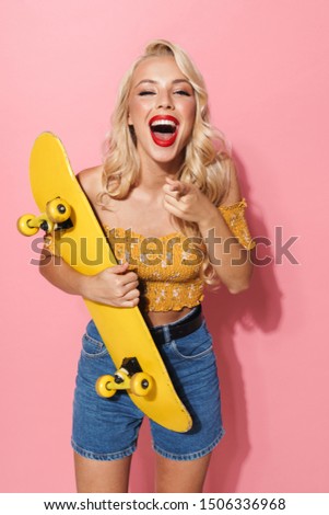 Image of fashion young woman with red lipstick wearing summer clothes smiling and holding skateboard isolated over pink background