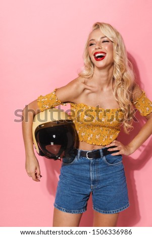 Image of gorgeous seductive moto girl wearing red lipstick smiling and holding motorbike helmet isolated over pink background
