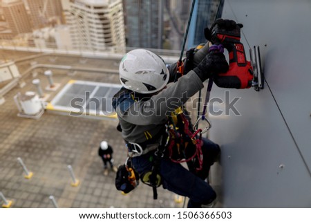 Top view of rope access abseiler wearing full body safety harness hard hat abseiling working at height wearing a safety hand glove protection using red electric hacksaw cutting aluminium wall panel 