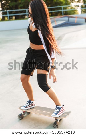 Photo of teenage young woman in streetwear wearing headphones riding skateboard in skate park
