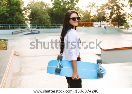 Photo of beautiful young woman in streetwear smiling while carrying skateboard in park