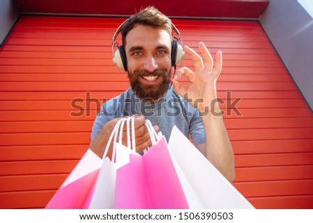 The best shopping. Portrait of bearded man with shock crazy emotion on his face with headphones looking at camera with ok sign with pink, white shopping bags. Outdoor shot on red wall background.