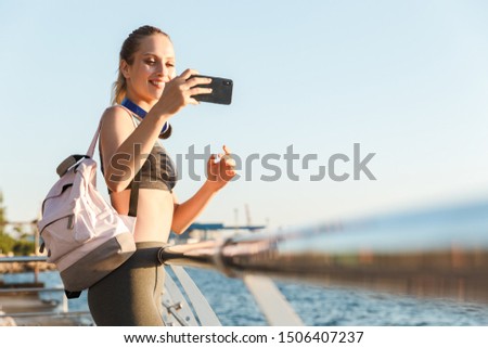 Beautiful young fitness woman carrying backpack walking at the pier, taking picture of a seascape on her phone