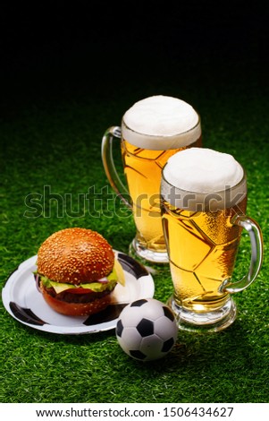 Two glasses of beer, hamburger and soccer ball on green grass. Table for soccer fans
