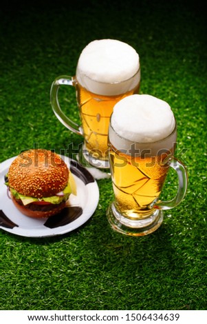 Two glasses of beer and hamburger on green grass. Table for soccer fans