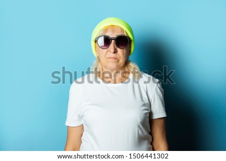 Funny old woman in a hat and glasses, makes a hand gesture on a blue background. Concept cool stylish grandmother, modern style