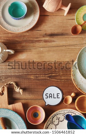 Top view of variety of colorful ceramic kitchen plates, bowls and cups on wooden table with 'sale' sign. Tableware sale concept with copyspace. 