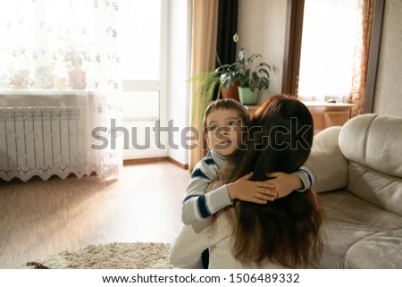 Mom hugs her son at home with light from the windows. Expression of family kindness and maternal love. Mutual understanding, sympathy and support of parents and children. Boy with neutral emotions. 