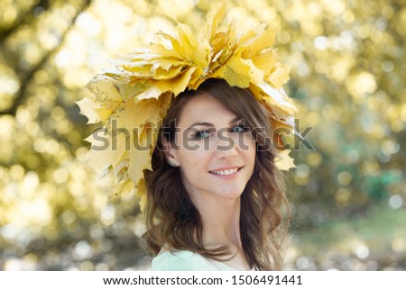 Closeup portrait of a beautiful smiling young brunette woman with a wreath of yellow maple leaves on her head. Golden autumn foliage of trees in the background