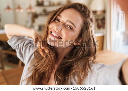 Picture of young smiling beautiful positive ginger woman indoors at home take selfie by camera.