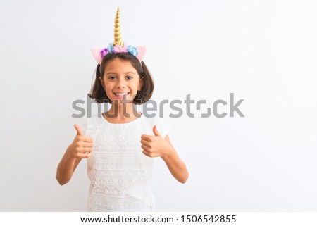 Beautiful child girl wearing unicorn diadem standing over isolated white background success sign doing positive gesture with hand, thumbs up smiling and happy. Cheerful expression and winner gesture.