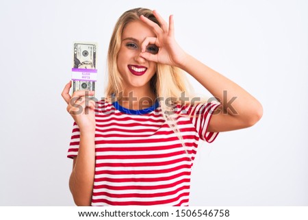 Beautiful woman wearing red striped t-shirt holding dollars over isolated white background with happy face smiling doing ok sign with hand on eye looking through fingers