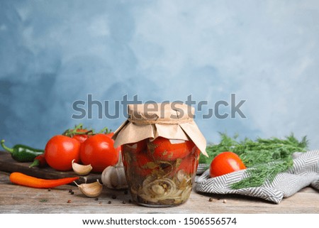Pickled tomatoes in glass jar and products on wooden table against blue background, space for text