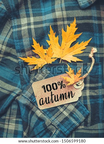 100% autumn - text on tag, autumnal maple leaves lying on blue plaid shirt, abstract background. autumn fashion style concept. fall season. Flat lay