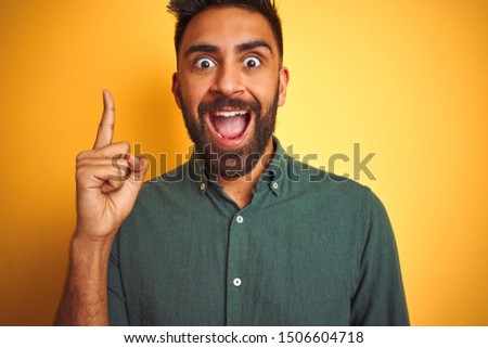 Young indian man wearing green shirt standing over isolated yellow background pointing finger up with successful idea. Exited and happy. Number one.