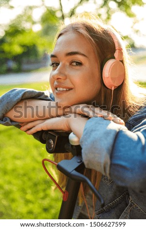 Image of a cheery smiling teenage girl outside in nature green park on grass listening music with headphones with scooter.