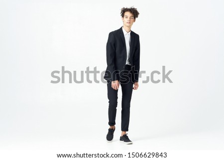 A guy with curly hair and a business suit is standing on a light, isolated background in full growth                           