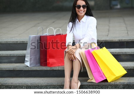 Portrait of cheerful female in stylish sunglasses looking at camera with happiness. Smiling woman sitting on stairs and putting hands on knees. Fashion and shopping concept. Blurred background