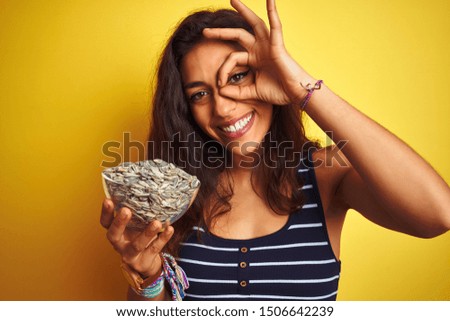 Young beautiful woman holding bowl with sunflowers seeds over isolated yellow background with happy face smiling doing ok sign with hand on eye looking through fingers