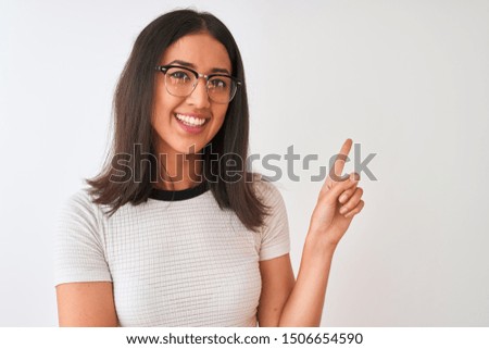 Chinese woman wearing casual t-shirt and glasses standing over isolated white background with a big smile on face, pointing with hand and finger to the side looking at the camera.