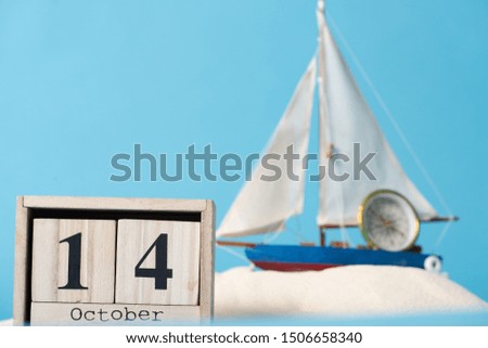 wooden calendar with 14 October date near decorative ship and compass  in white sand isolated on blue