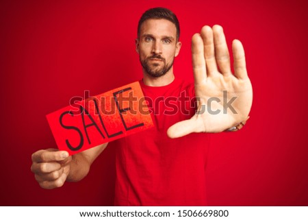 Young man holding sale advertising poster board over red isolated background with open hand doing stop sign with serious and confident expression, defense gesture
