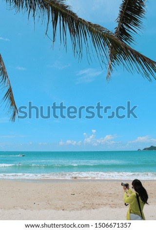 An unrecognizable girl photographer shoots on the beach under the coconut trees and has a speedboat sailing on the surface. The background is the exotic tropical coastline of Phuket, Thailand.