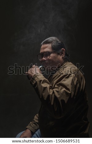 A  man wearing dark green jacket and glasses with e-cigarette Vape mode and vaping. Low light, grey background.