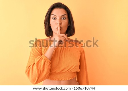 Young beautiful woman wearing casual t-shirt standing over isolated orange background asking to be quiet with finger on lips. Silence and secret concept.