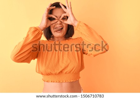 Young beautiful woman wearing casual t-shirt standing over isolated orange background doing ok gesture like binoculars sticking tongue out, eyes looking through fingers. Crazy expression.