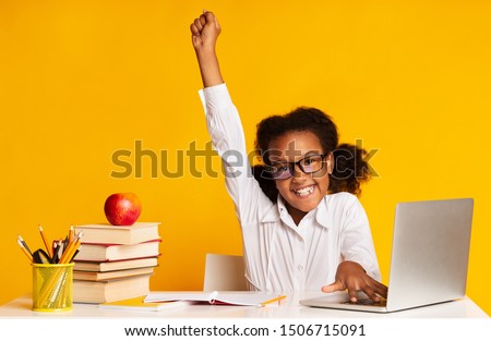 Black Elementary Student Girl Raising Hand Sitting At Laptop Doing Homework Over Yellow Background. Studio Shot Royalty-Free Stock Photo #1506715091