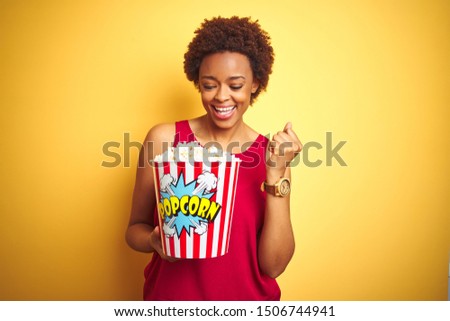 African american woman holding pack of popcorn over yellow isolated background screaming proud and celebrating victory and success very excited, cheering emotion