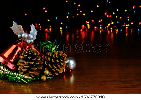 Pine cones, red Christmas bells, ornaments, silver bauble, green tinsel garland for Christmas decoration on wooden table with bokeh multicolored light background.