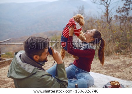 Gorgeous smiling Caucasian brunette sitting on blanket and posing with her dog while her boyfriend taking picture of them. Picnic at autumn concept.