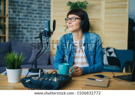 Beautiful young woman blogger in headphones is recording podcast using microphone sitting at table at home. Blogging, lifestyle and audio records concept.