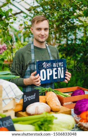 Cheerful farmer is holding we are open sign working in farm market selling organic food inviting people to harvest sale. Small business and shopping concept.