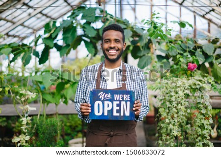African American man is standing in greenhouse with we are open sign smiling looking at camera welcoming buyers for fresh flowers. Business and nature concept.