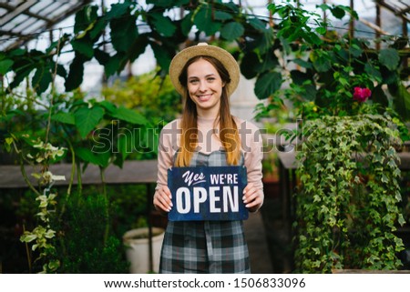 Beautiful young lady wearing hat and apron is standing in greenhouse with we are open sign smiling welcoming buyers. Business and floristry concept.